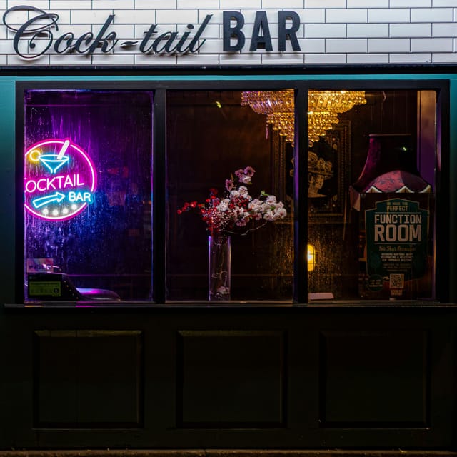 A bar front at night, its window lit by a pink neon cocktail sign, a vase of blossom and a chandelier