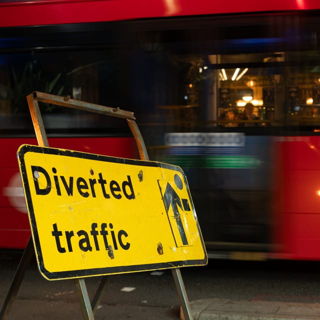 A yellow Diverted traffic sign on a pavement at night, a red London bus blurring past behind it